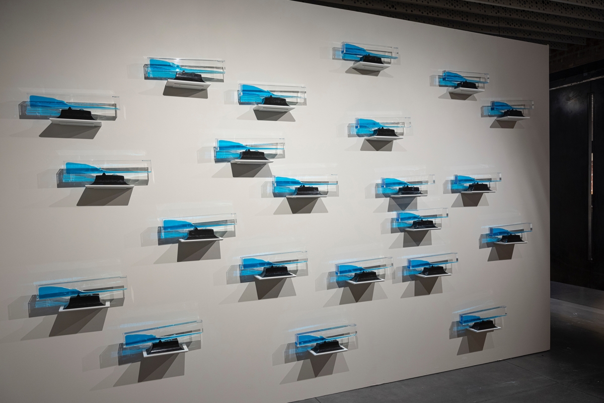 Gallery wall installation featuring multiple small, identical sculptural pieces with blue and clear components mounted in a grid-like arrangement.