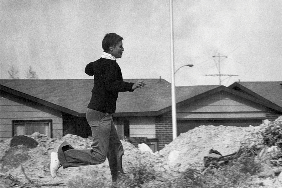 Black-and-white photograph of a person running across a dirt or construction area with houses visible in the background.