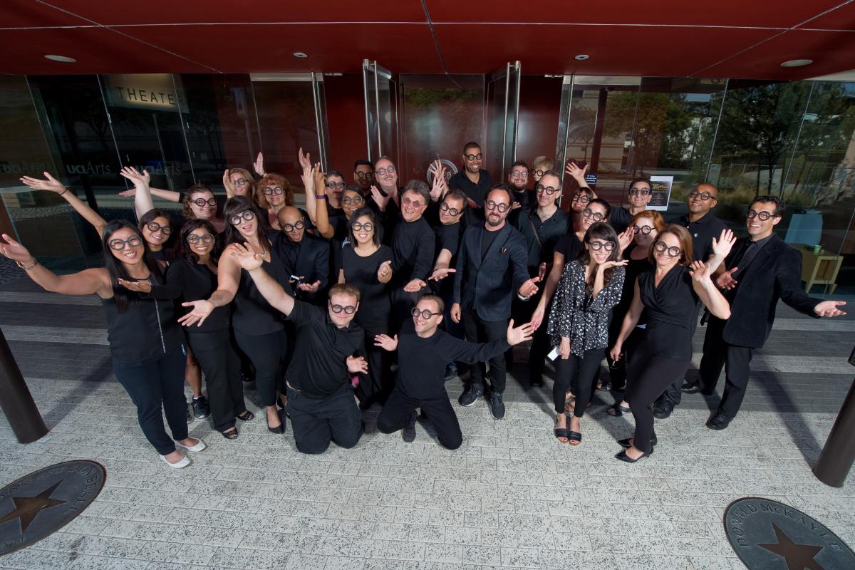 Large group of people dressed in black posing together outside a building entrance, arranged in rows and smiling at the camera.