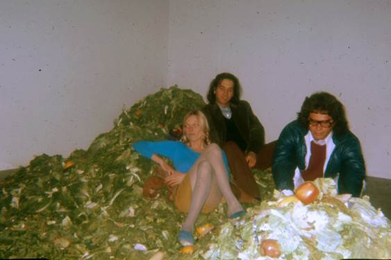 Three people seated on the floor beside a large pile of organic debris or compost material, including leaves and discarded food items, inside a plain room.