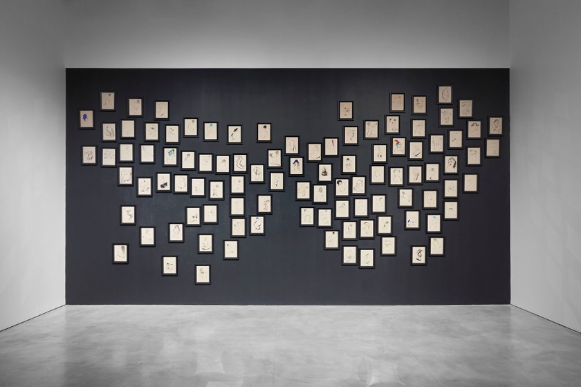 Gallery wall displaying many small framed drawings arranged in a clustered, map-like formation against a dark background wall.