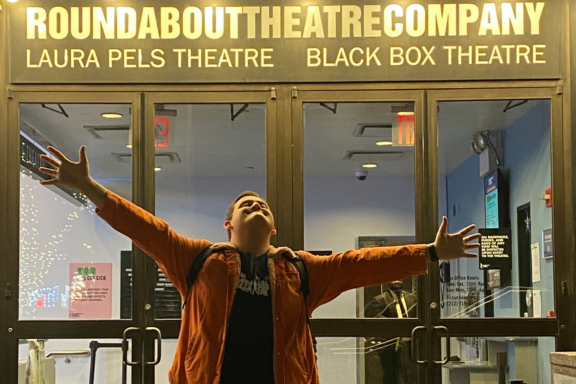 Person standing outside glass theater doors with arms outstretched, beneath signage reading “ROUNDABOUT THEATRE COMPANY,” “LAURA PELS THEATRE,” and “BLACK BOX THEATRE.”