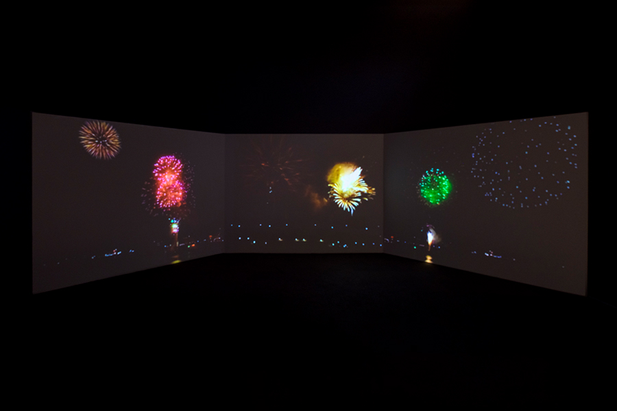A dark gallery space features a wide, multi-panel projection of colorful fireworks bursting across three connected screens. Bright red, yellow, and green explosions illuminate the room against a black background, creating an immersive visual display.