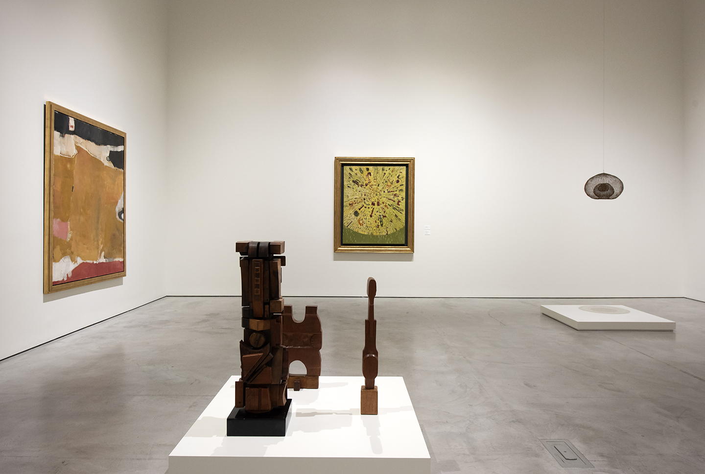 A spacious gallery with white walls and polished concrete floors displays a mix of paintings and sculptures. Two wooden sculptures sit on a white pedestal in the foreground, while framed artworks hang on the surrounding walls, including a large abstract painting and a smaller piece centered on the far wall.