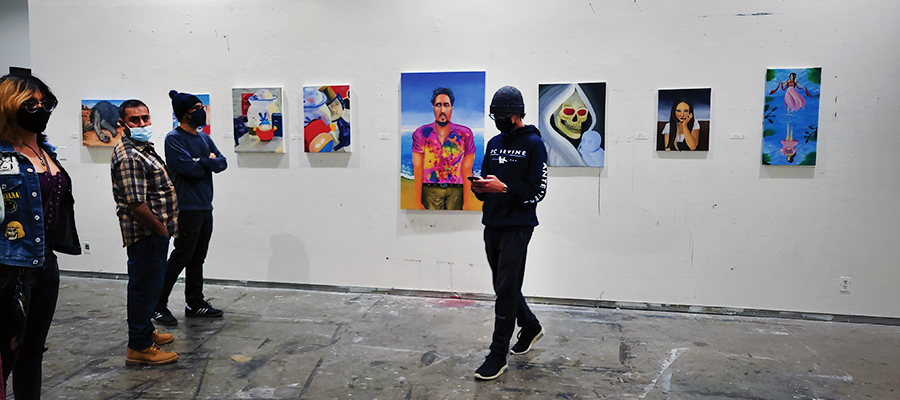 In a gallery space with concrete floors, several people wearing face masks stand and talk while viewing colorful portrait paintings displayed on a white wall. One person walks across the room in the foreground as others observe the artwork.