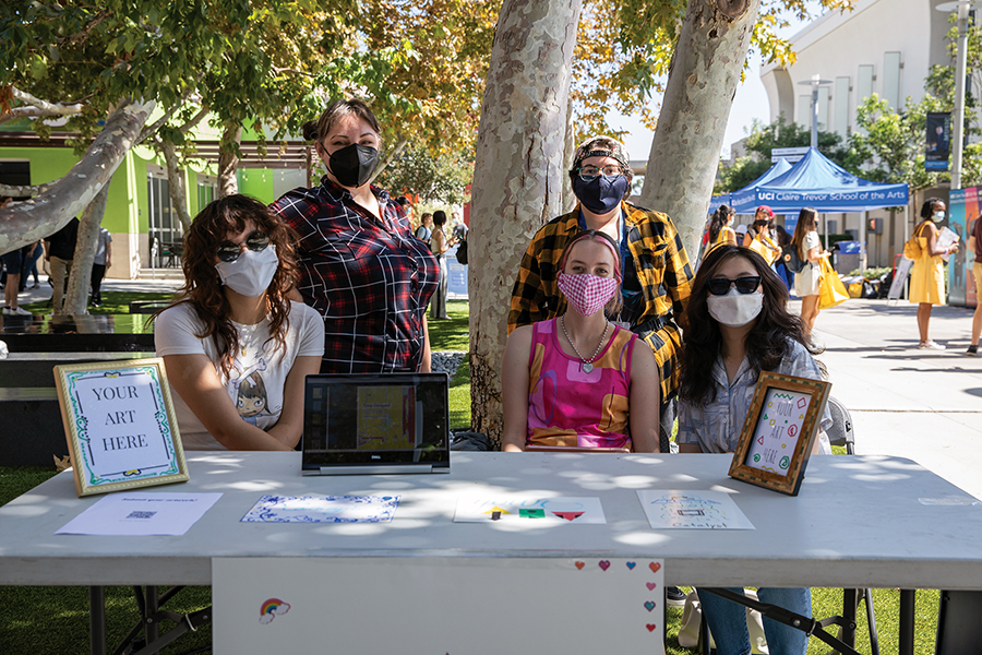 Five people wearing face masks pose behind an outdoor information table on a sunny campus walkway. The table displays small artworks, a tablet, and a sign that reads “Your Art Here,” while tents and other event booths appear in the background beneath leafy trees.