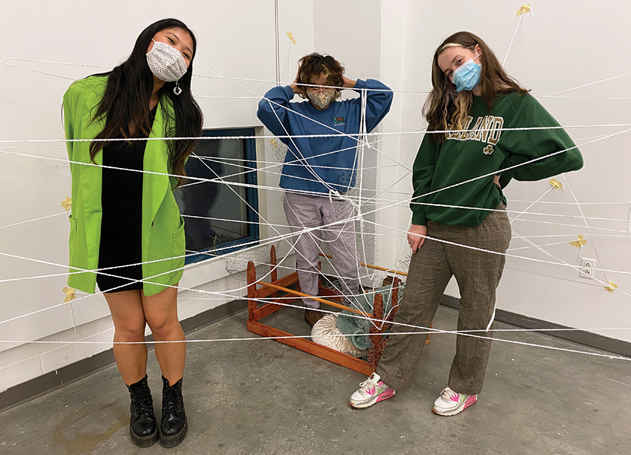 Three people wearing face masks stand inside a gallery space, entangled in a web of white string stretched across the room. A small wooden structure with additional string sits on the floor behind them, suggesting an interactive art installation.