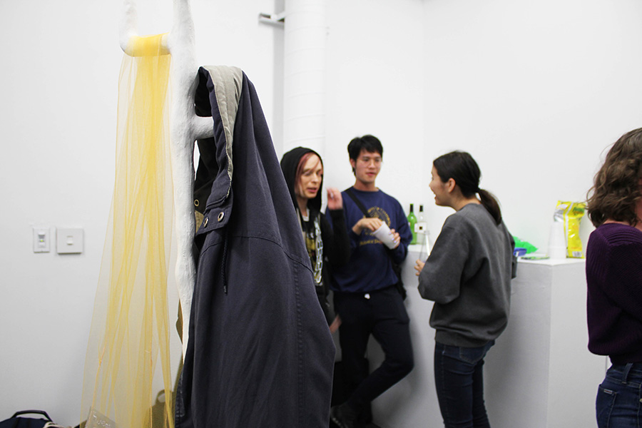 In a white gallery space, several people stand and talk near a pedestal. In the foreground, a coat hangs over a tall object beside a sheer yellow fabric drape, partially obscuring the view of the group conversing in the background.