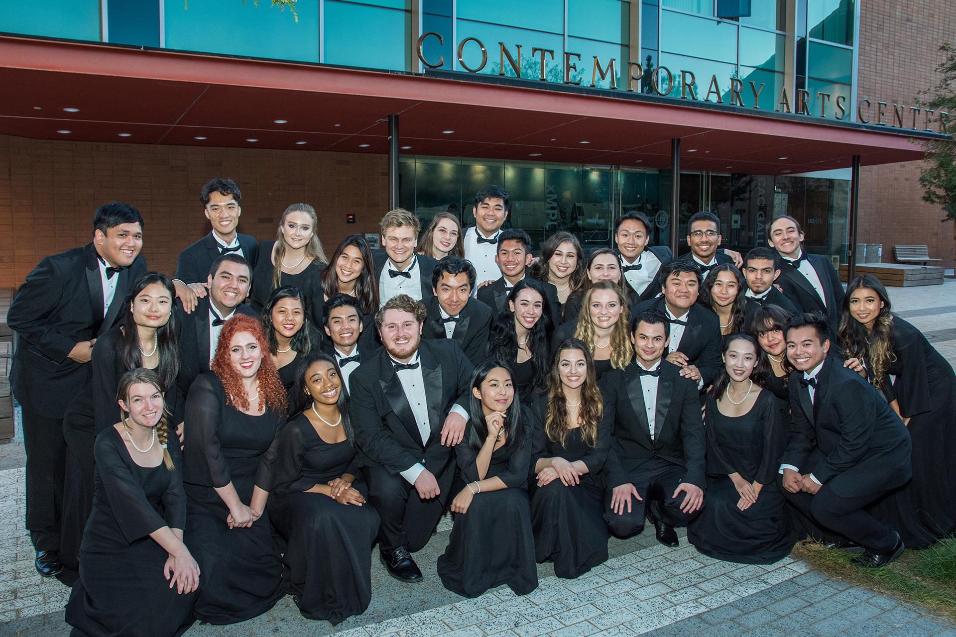 A large group of students dressed in formal black attire poses together outside the Contemporary Arts Center at dusk. They stand and kneel in rows in front of the building’s glass entrance and red overhang, smiling for a group photo.