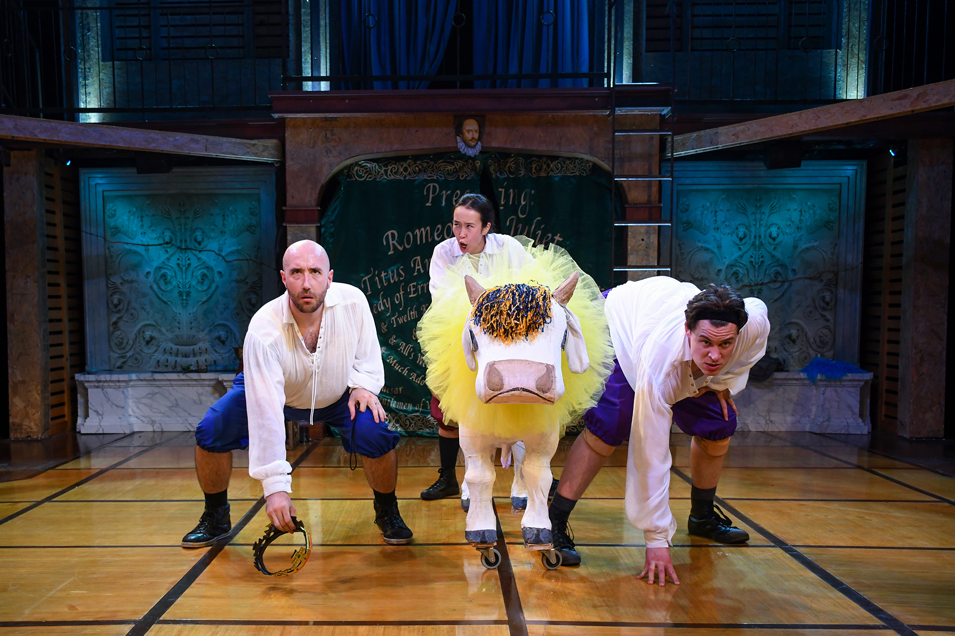 Three performers in period-style costumes crouch on a stage floor, holding props, while a large, whimsical bull prop with yellow fur stands between them. A chalkboard backdrop and theatrical set pieces frame the scene under stage lighting.