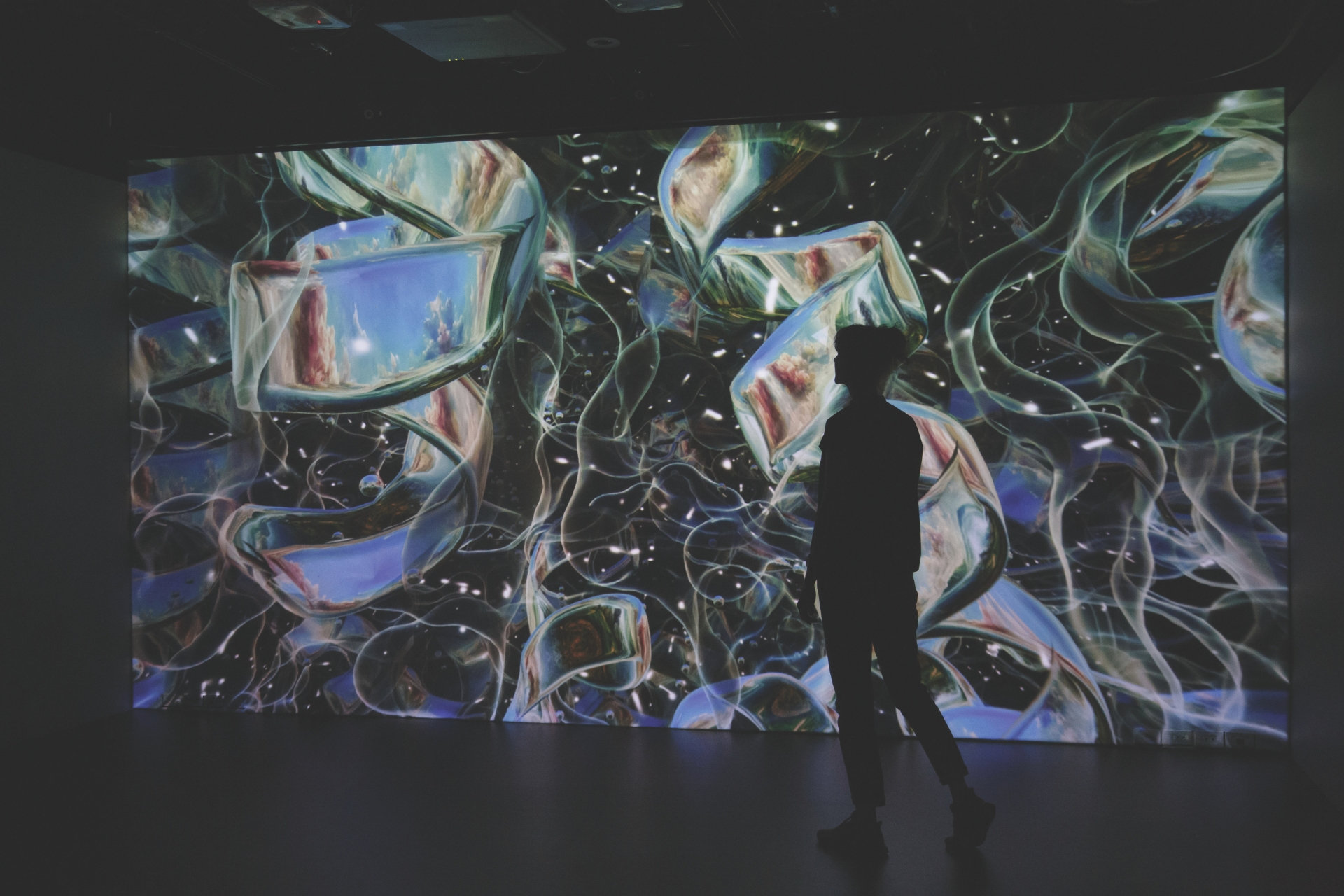A silhouetted figure stands in front of a large immersive projection covering the wall. The digital artwork displays abstract, swirling imagery with floating shapes and web-like lines in shades of blue, green, and white, filling the dark gallery space with light.