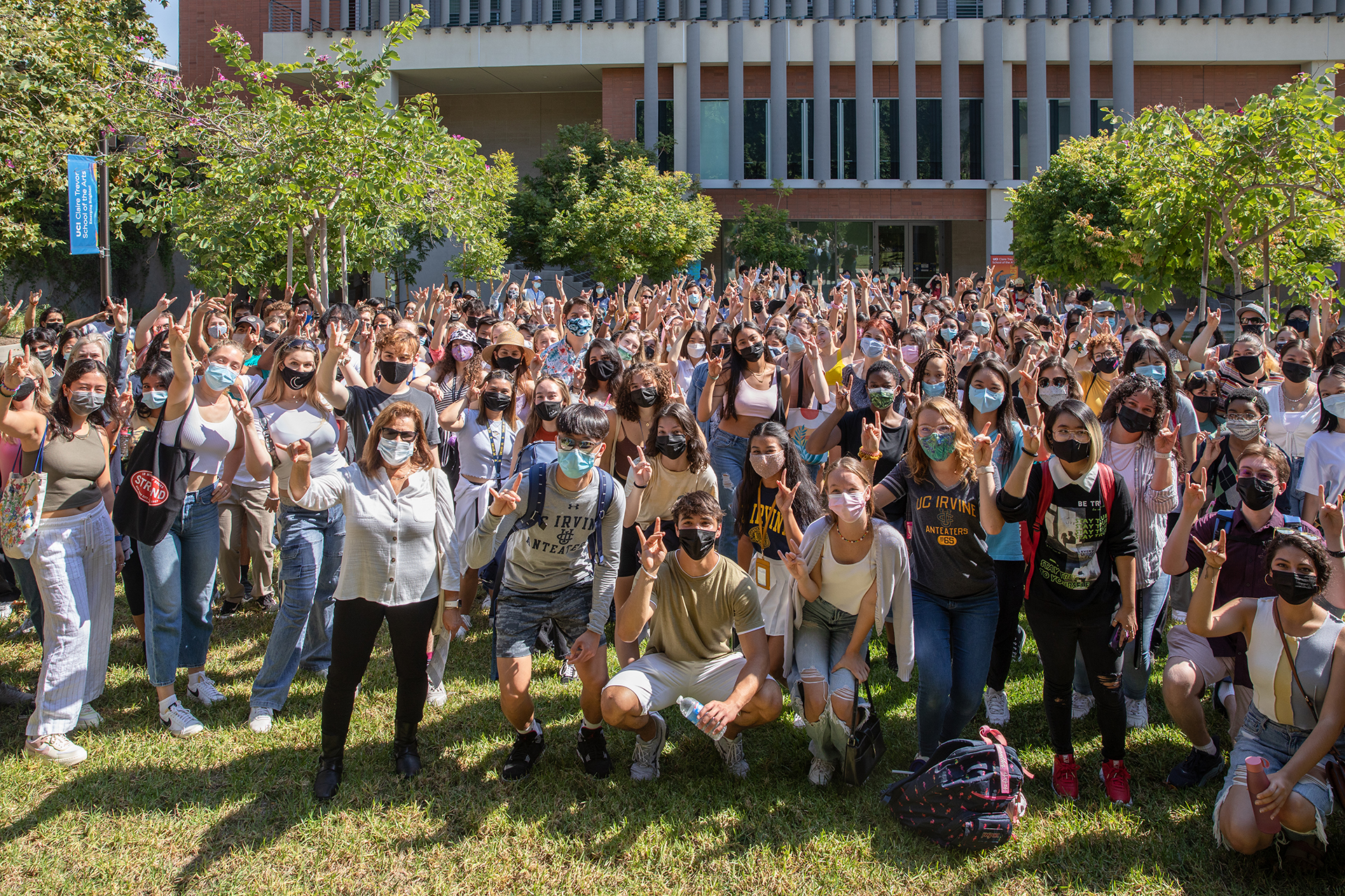 A large crowd of students gathers outdoors on a grassy lawn in front of a campus building. Many wear face masks and raise their hands or wave toward the camera, posing together in a lively group photo under sunny skies.