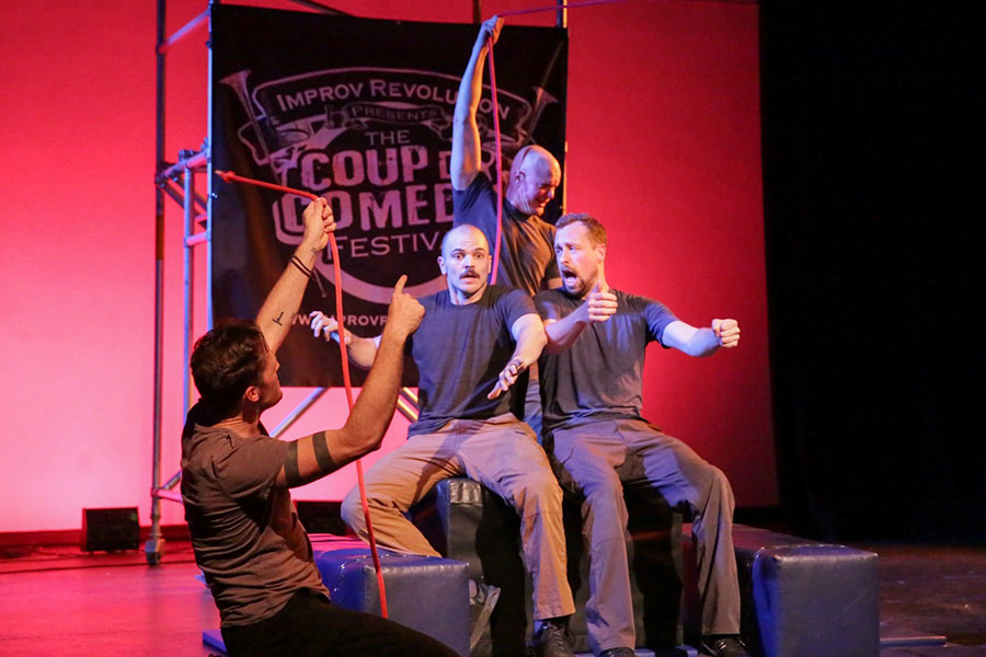 Four performers act out a comedic scene onstage under red lighting. Three sit on a large prop while another stands behind them holding ropes, with a banner in the background reading “Improv Revolution: The Coup de Comedy Festival.”
