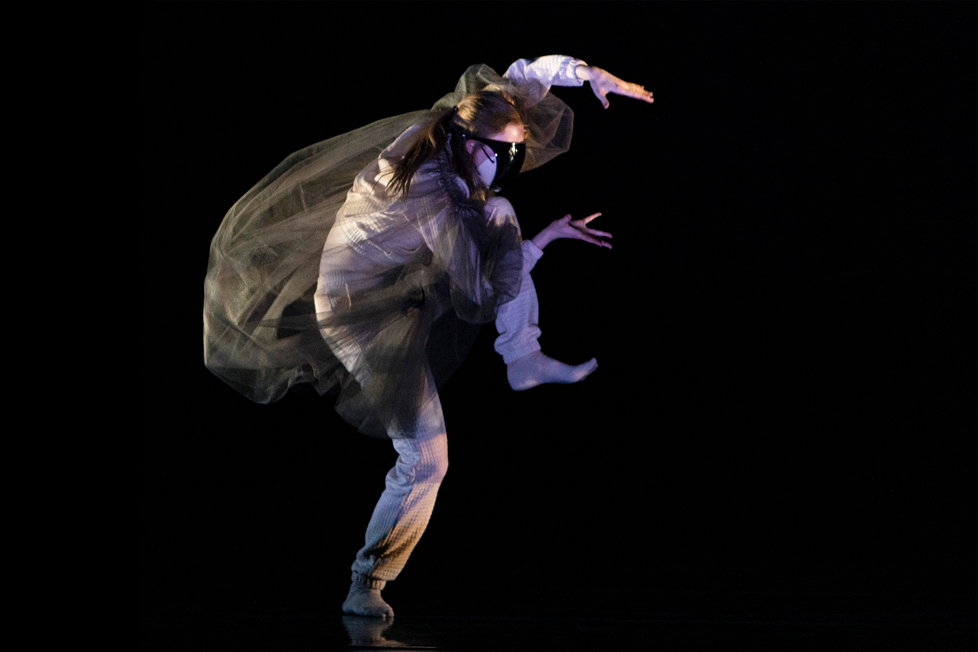 A dancer performs on a dark stage, captured mid-motion with one leg lifted and arms curved overhead. Wearing layered, flowing fabric that billows around the body, the performer is illuminated by focused stage lighting against a black background.