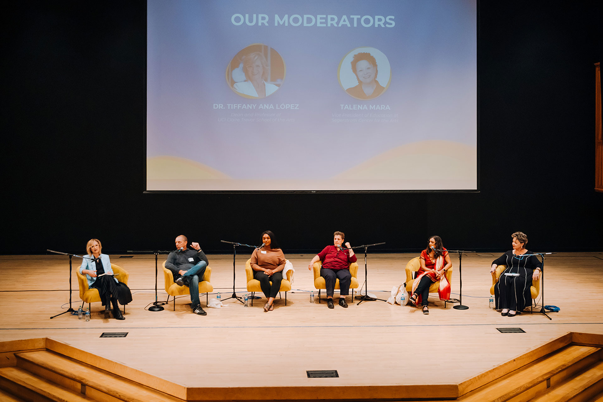 A panel of six speakers sits onstage in armchairs with microphones, participating in a discussion. A large screen behind them displays the heading “Our Moderators” along with two profile images, while the audience area is out of view.
