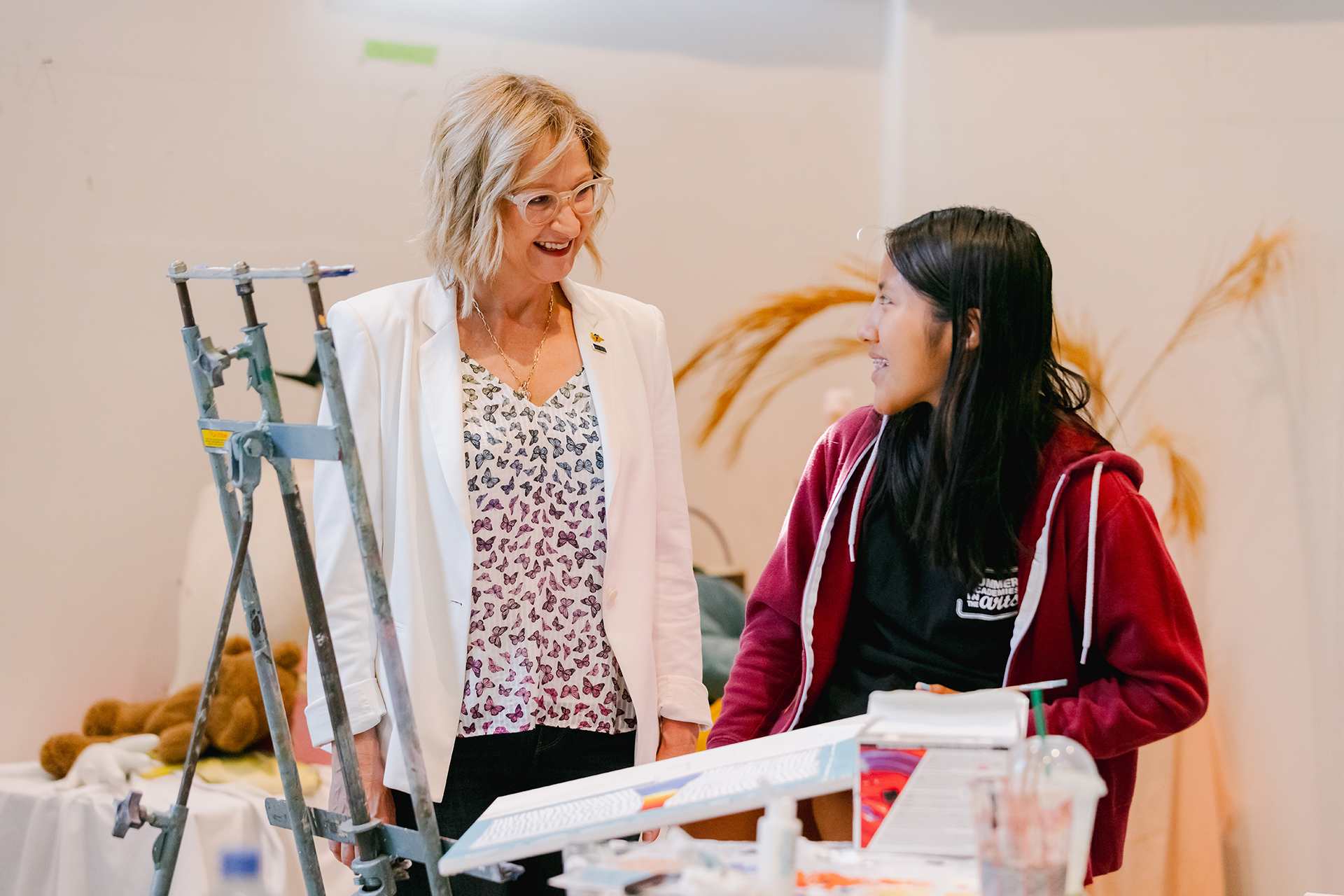 Two women stand together in an art studio, smiling and talking beside an easel and drawing board. One wears a white blazer and glasses, while the other wears a red hoodie, as they look at each other in conversation.