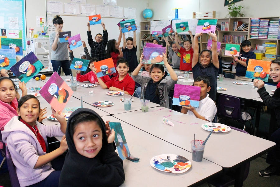 A classroom of children smiles and holds up colorful paintings they have created, each featuring a bright sunset scene. Paint palettes and cups of water sit on the tables, while a teacher stands at the back of the room.