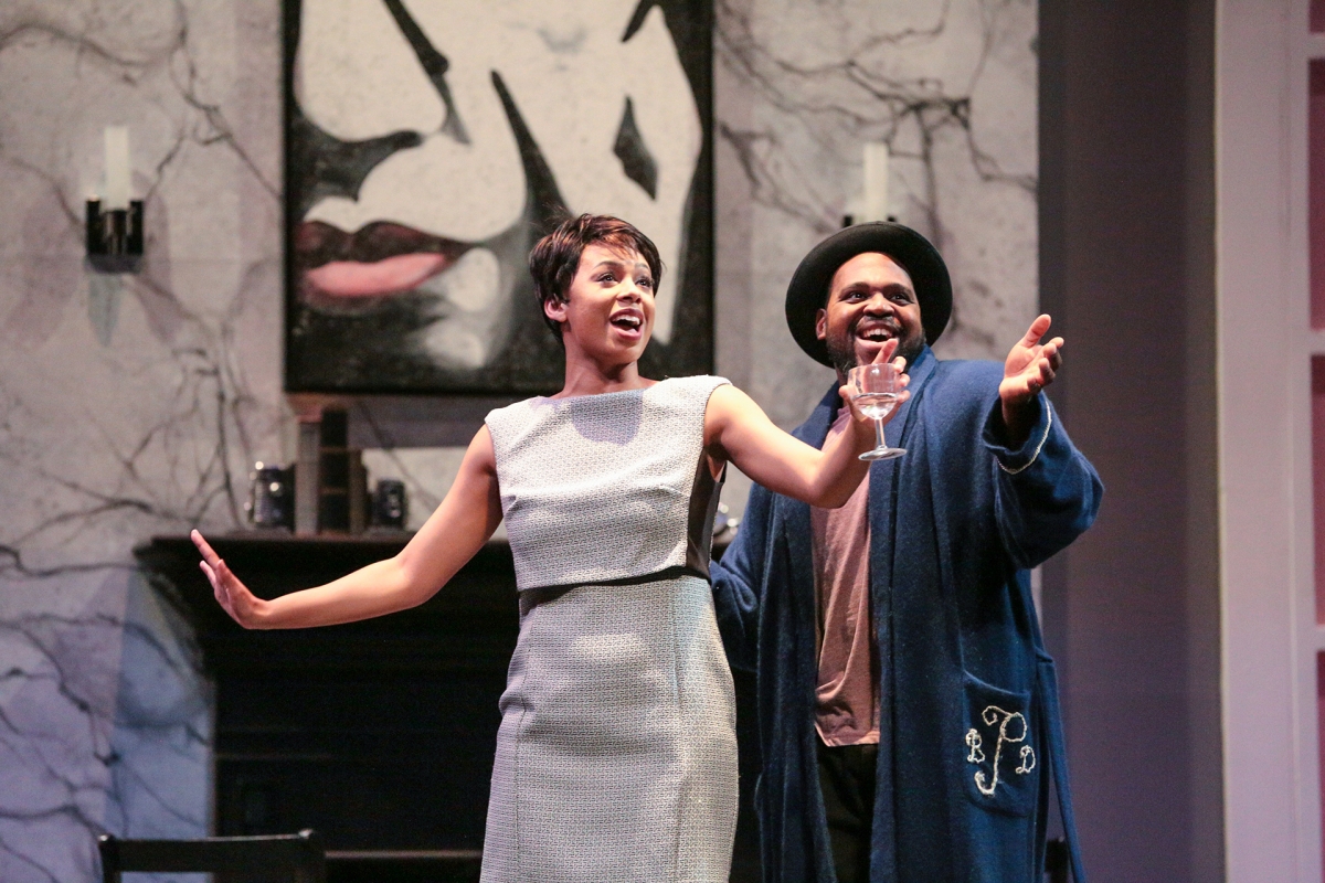 Two performers stand onstage in a living room–style set, smiling and gesturing outward as if addressing an audience. One wears a light-colored dress while the other wears a robe and hat, holding a glass, with a large portrait painting visible behind them.