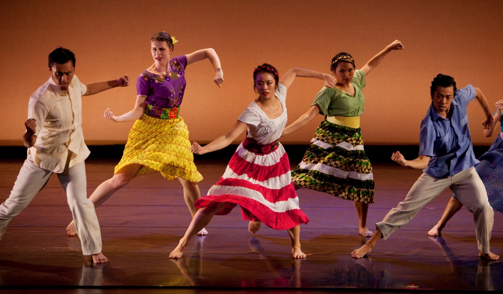 Five dancers perform onstage against a warm orange backdrop, wearing colorful skirts and tops. They move in a lively, synchronized pose with bent arms and extended legs, captured mid-step under stage lighting.