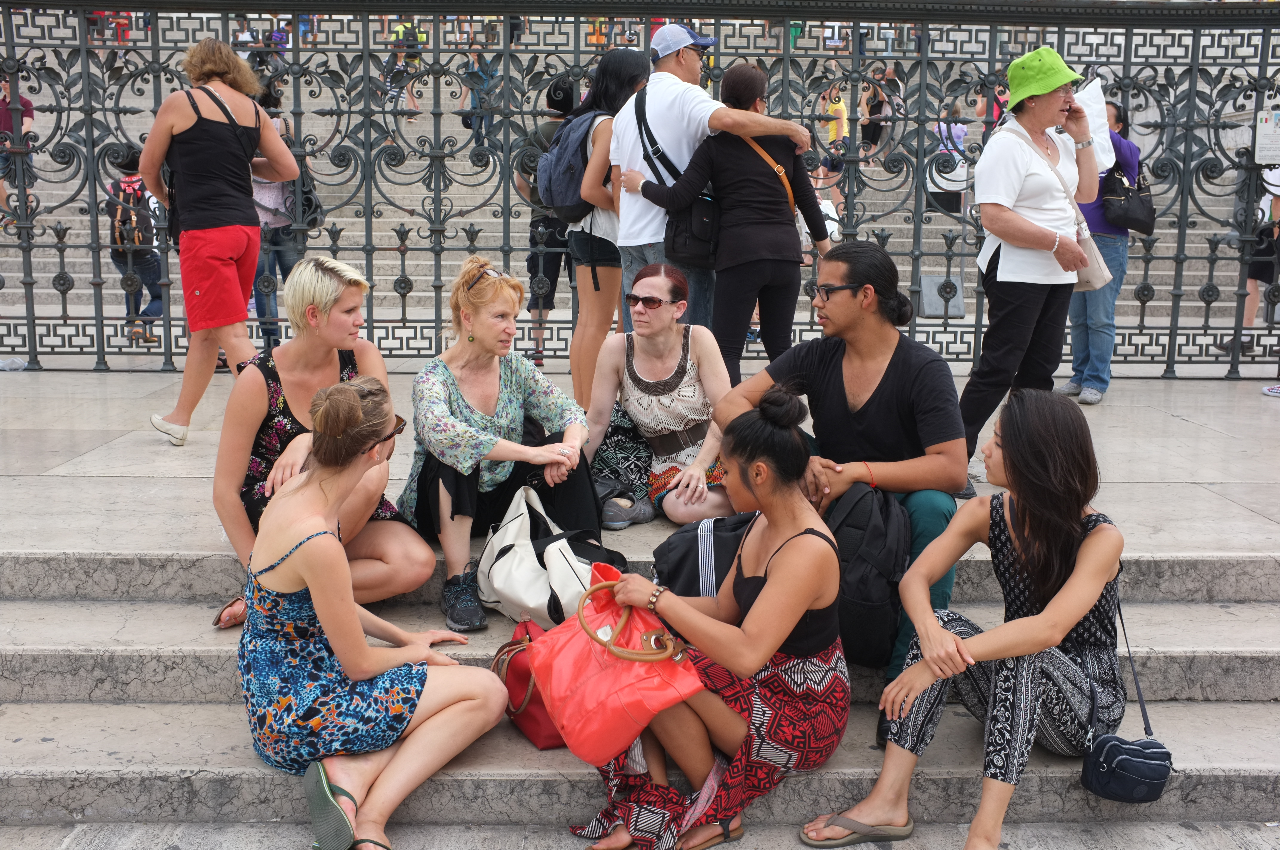 A group of people sits together on stone steps, talking and resting in front of a decorative metal fence. Some hold bags and personal items, including a bright red tote placed in the center. Other pedestrians move in the background.