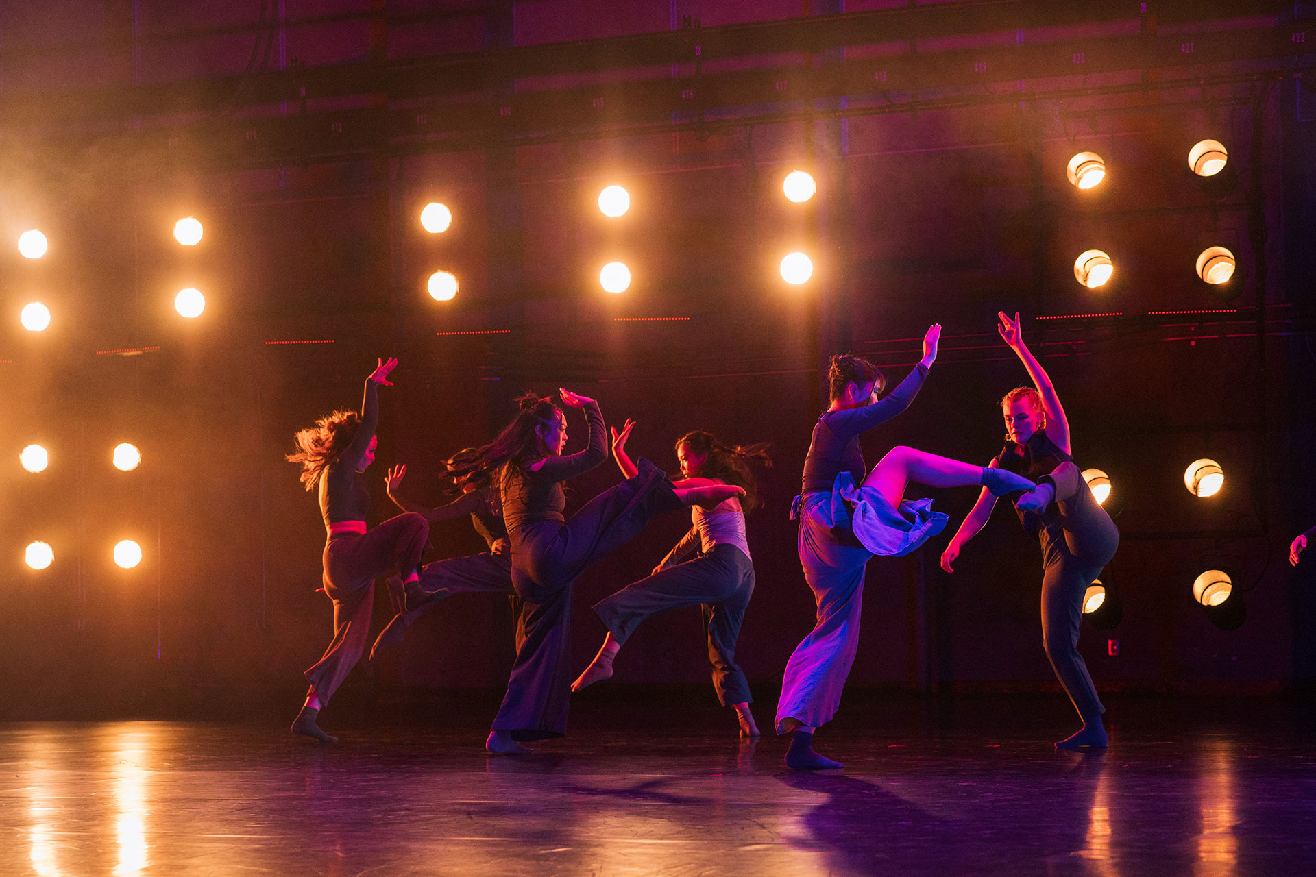 A group of dancers performs onstage under bright amber stage lights. They move dynamically in pairs and small clusters, with lifted legs and raised arms silhouetted against the glowing backdrop.