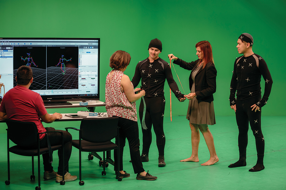 In a green screen studio, several people work with performers wearing motion capture suits marked with tracking points. A large monitor displays a 3D skeletal animation of movement, while one person measures or adjusts a performer’s arm as others observe.