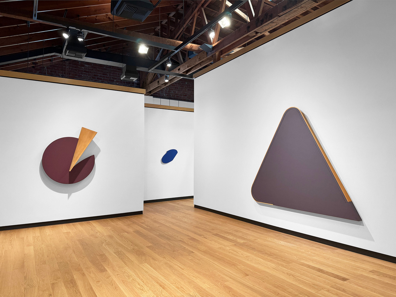 A gallery room with white walls and a light wood floor displays large geometric wall-mounted artworks. On the left wall, a circular maroon shape is partially overlaid with a yellow triangular form. In the center background, a small blue oval piece is mounted. On the right wall, a large triangular form in muted purple with a narrow wooden edge detail is displayed. Overhead track lighting illuminates the space, and exposed wooden beams are visible on the ceiling.