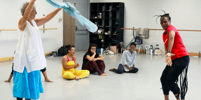 Two dancers move energetically in a studio, one lifting flowing fabric and the other stepping forward expressively, while several people sit on the floor watching in the background among mirrors and ballet barres.