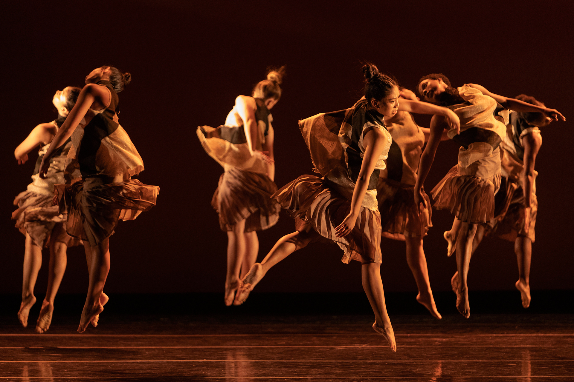 Several dancers in flowing, earth-toned costumes leap across a stage under warm amber lighting. Their skirts flare midair as they move in unison against a deep red backdrop.