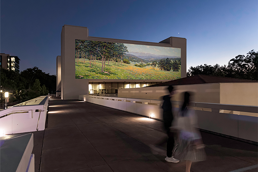 A person walks past a large outdoor projection of a mountain landscape displayed on a concrete wall at night, with a lit multi-story building visible in the background.