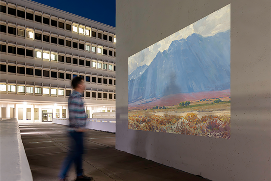 A person walks past a large outdoor projection of a mountain landscape displayed on a concrete wall at night, with a lit multi-story building visible in the background.