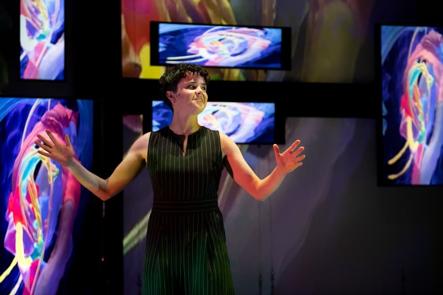 Person in pinstriped dress performs in front of multiple screens displaying colorful abstract art.