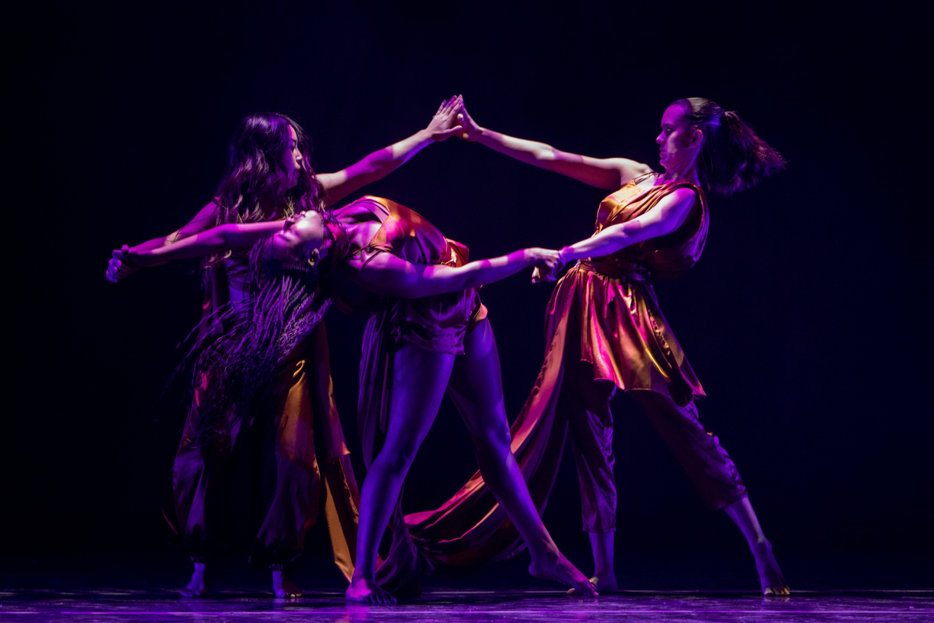 Three dancers perform on stage under purple lighting with arms extended and hands touching in a connected pose.