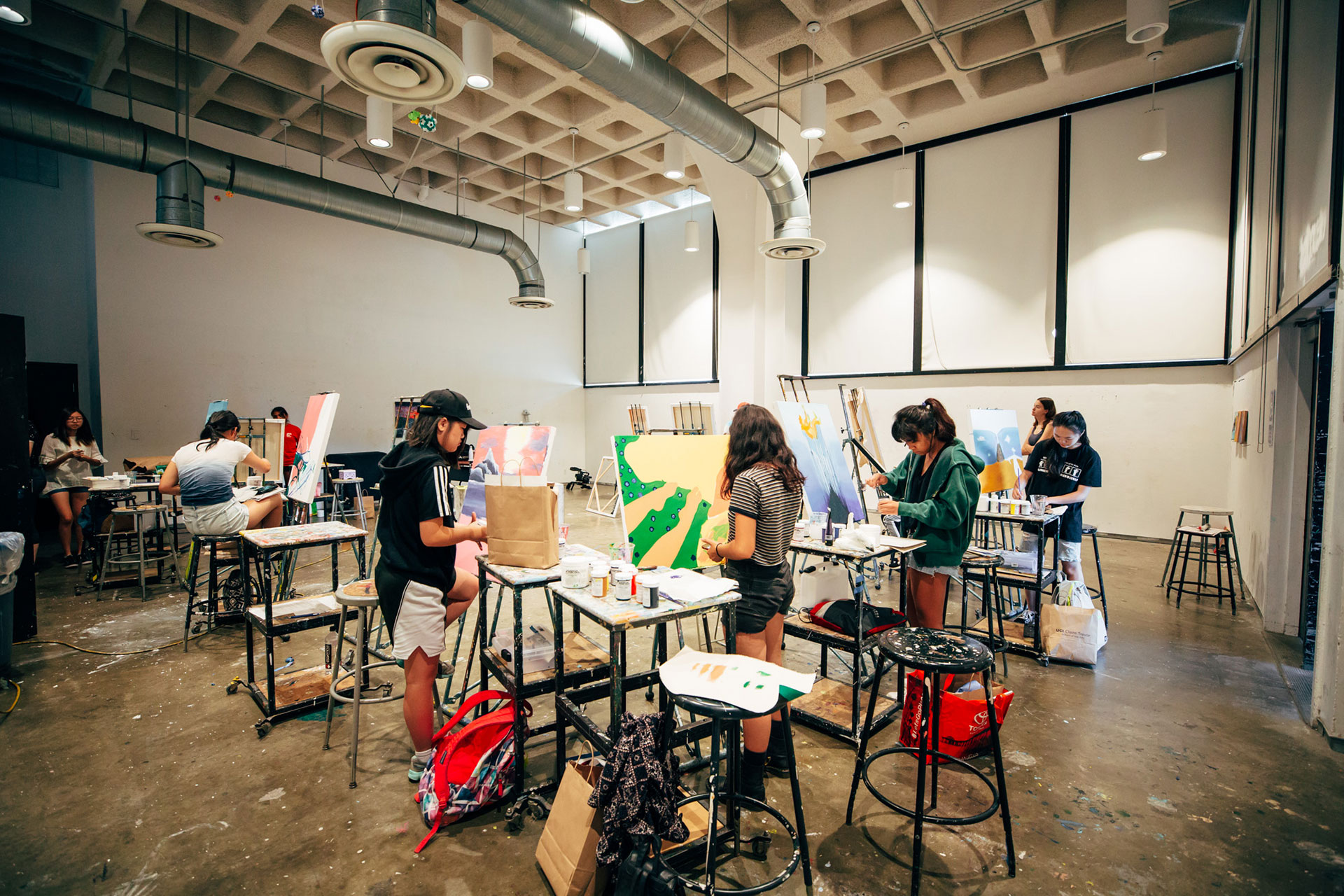 Students stand at easels painting in a large art studio with high ceilings, stools, and supplies spread across tables.