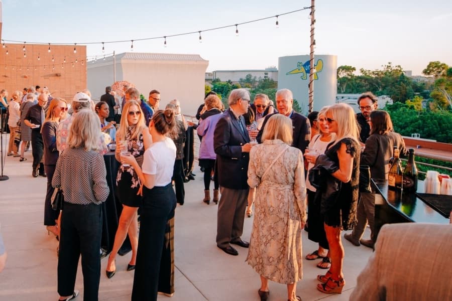 A diverse group of people socializes at an outdoor UCI rooftop reception under string lights.