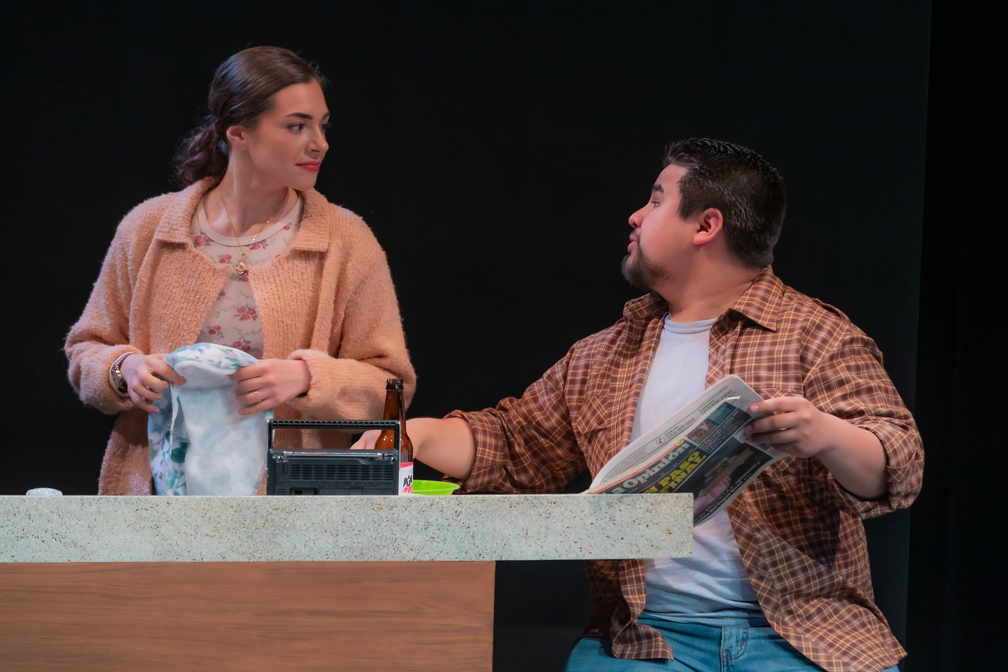 Two actors stand behind a kitchen-style counter on a stage set. One person in a tan jacket holds a dish towel and looks to the side at the other actor, who wears a brown plaid shirt over a white T-shirt and holds a folded newspaper. A small radio and kitchen items sit on the countertop, and the dark stage background isolates the pair under warm lighting.