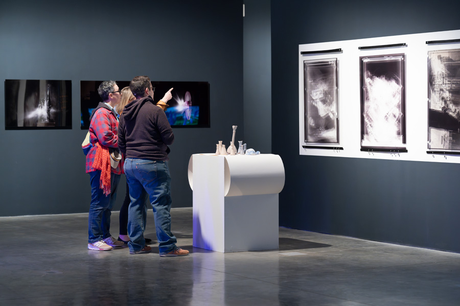 Three people stand in a gallery looking at photographs displayed on dark walls. One person points toward a screen mounted on the wall. A white pedestal in the center holds several small objects, and framed black-and-white images hang on the wall to the right.