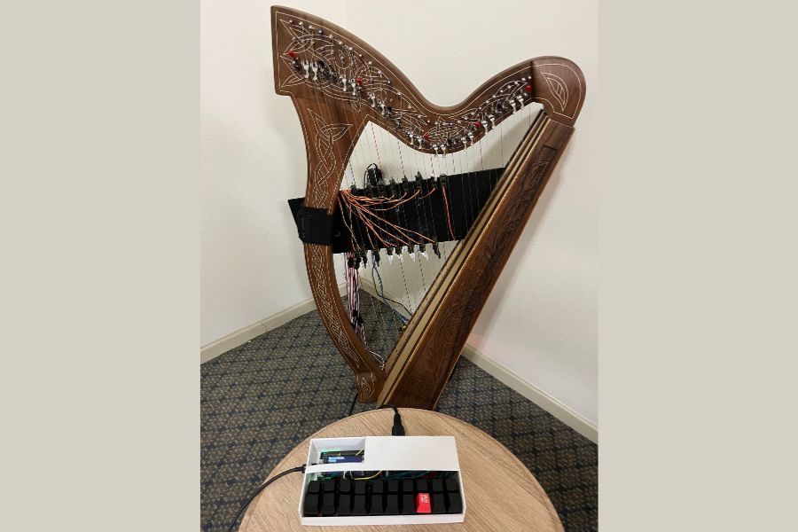 Wooden harp with carved detailing stands in the corner of a room. A small electronic device with buttons rests on a round table in the foreground, connected by a cable.