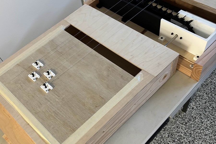 Close-up of a wooden instrument component resting on a table, showing layered plywood construction, small white brackets mounted on the surface, and strings visible from the adjacent structure.