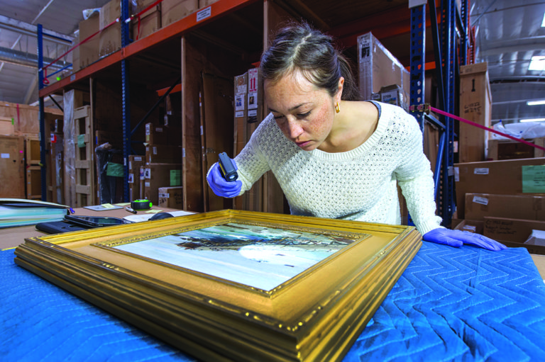 A person wearing blue gloves examines a framed artwork on a padded table while holding a small handheld tool close to the surface. Shelves filled with boxes and storage items line the warehouse space behind them.