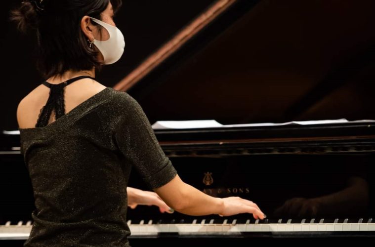 A musician wearing a face mask sits at a grand piano, viewed from behind and slightly to the side. Their hands rest on the keyboard while sheet music lies open on the piano’s music stand. The polished black piano reflects stage lighting.