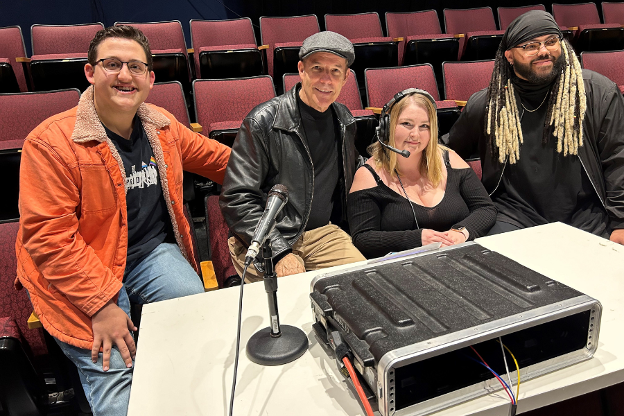 Four people sit together behind a table in an empty theater, smiling toward the camera, with microphones and audio equipment set up in front of them and rows of red auditorium seats in the background.