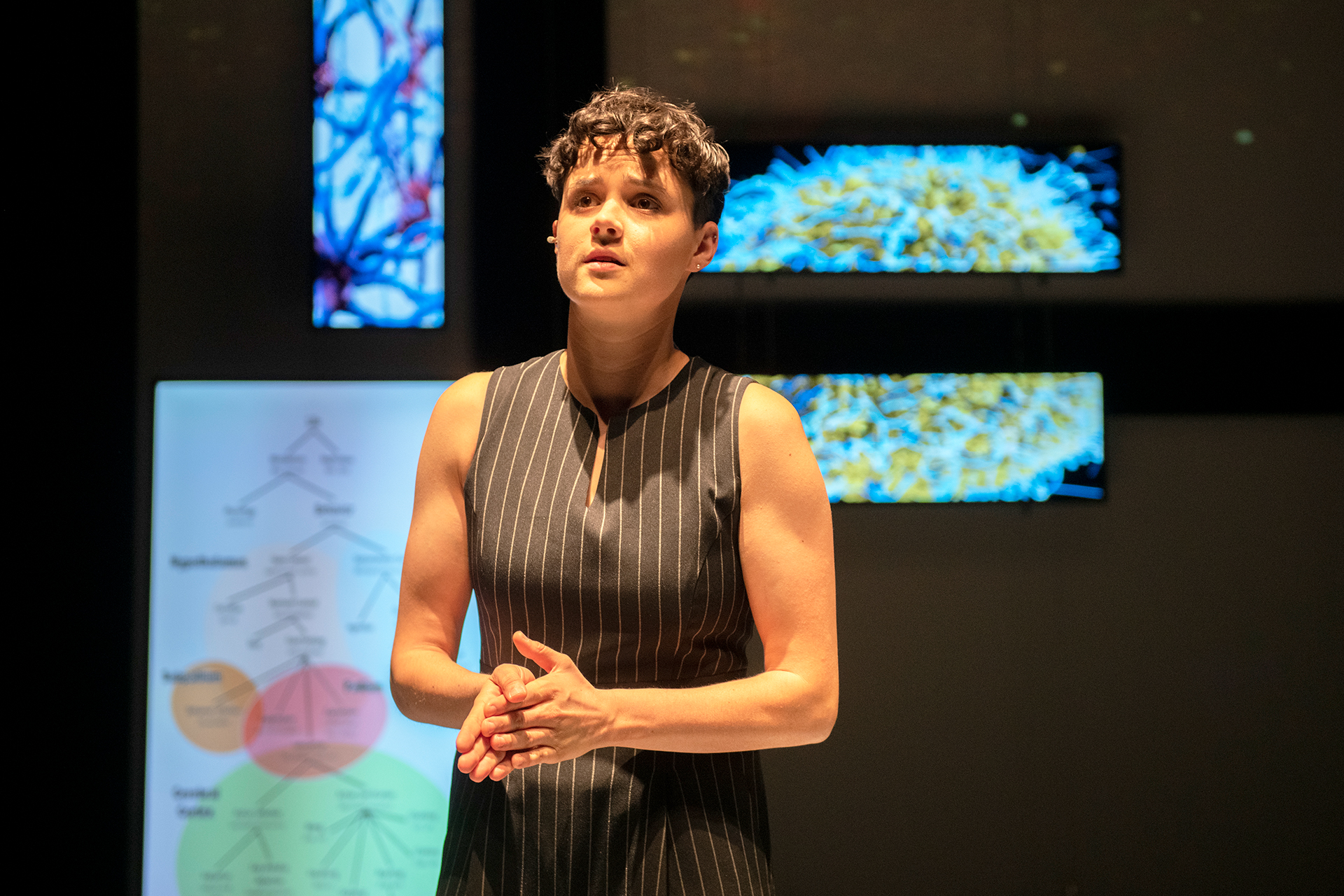 A person stands on a stage with hands clasped together, wearing a sleeveless black pinstriped outfit. Behind them, illuminated screens display colorful diagrams and abstract images. Warm stage lighting highlights the speaker against the darker background.