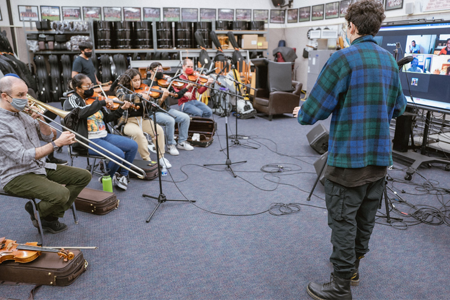 A small group of musicians rehearses in a room filled with instruments and equipment. String players sit with violins and violas, reading from music stands while microphones are positioned in front of them. A conductor stands facing the ensemble with arms raised, guiding the performance, while posters and shelves line the walls behind them.