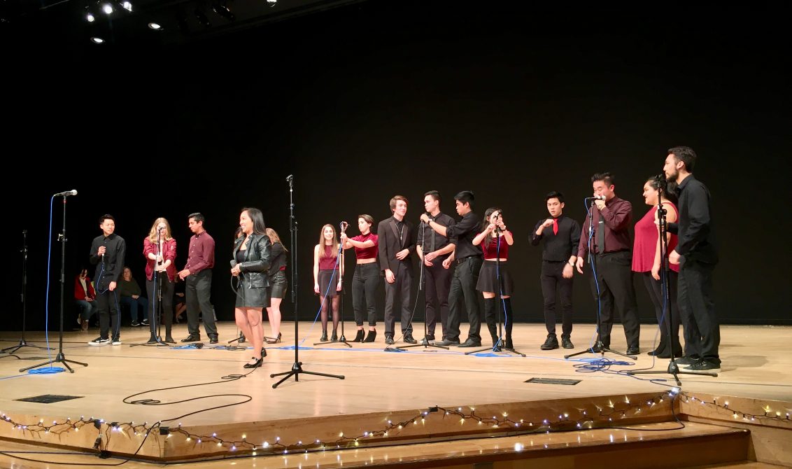 A group of performers stands across a stage in a theater, dressed mostly in black with red accents. Several hold microphones while others stand near mic stands, appearing to sing or prepare to perform. String lights line the front edge of the stage, and the background is a dark auditorium setting with overhead stage lighting.