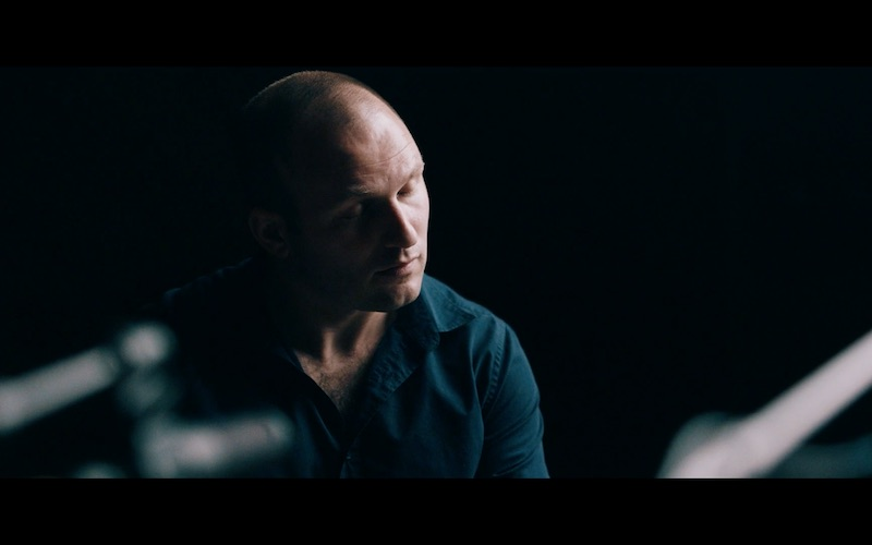 Moody, low-light portrait of a bald man in a dark shirt looking downward in a dim setting, with soft highlights on his face and a blurred foreground element suggesting a table or instrument.