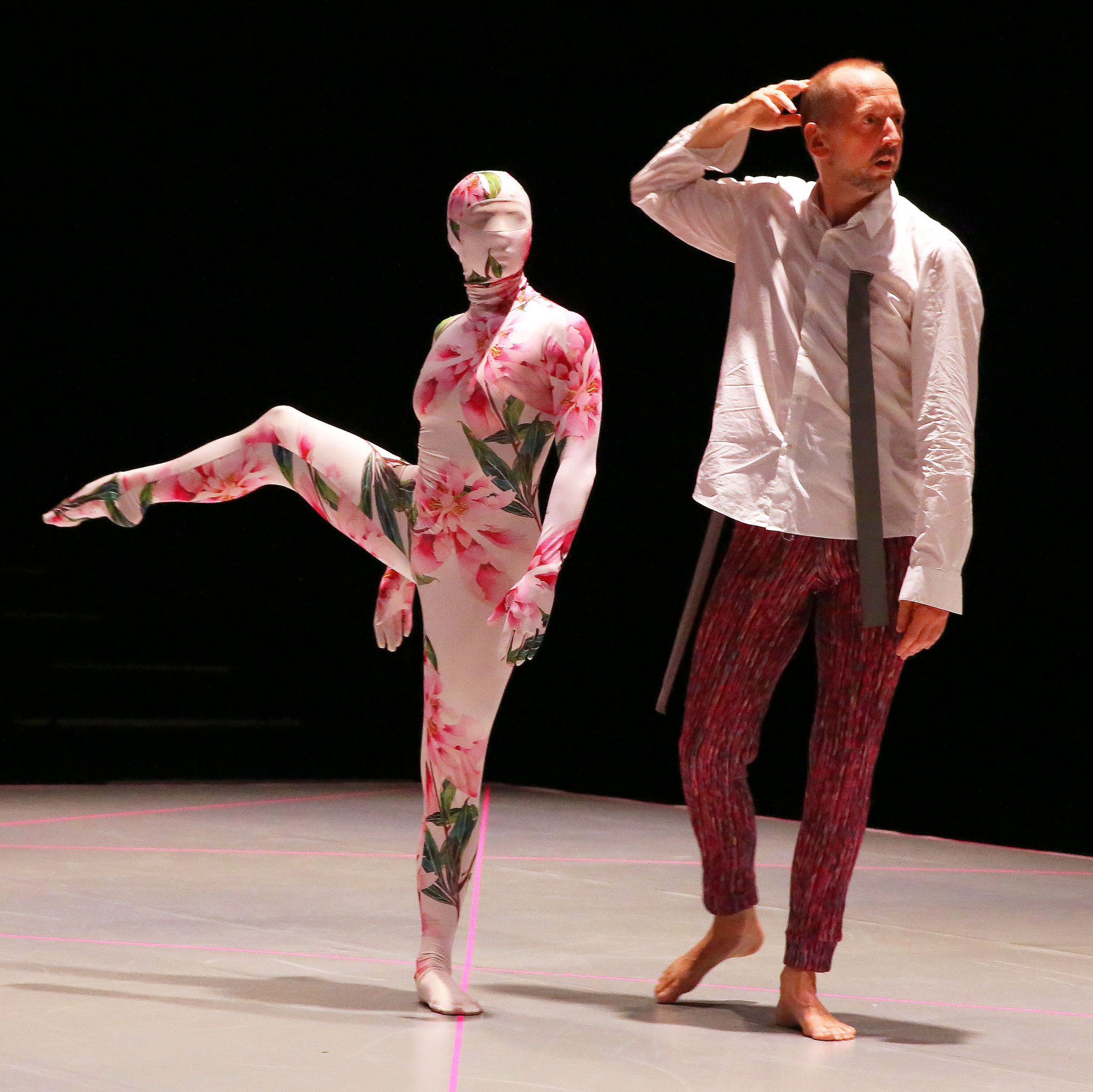 Two performers stand on a stage against a dark background. One performer wears a full-body suit with a floral pattern that covers the face and body, balancing on one leg with the other lifted. The other performer, dressed in a white shirt, loose tie, and patterned pants, stands barefoot with a hand raised to the side of his head.