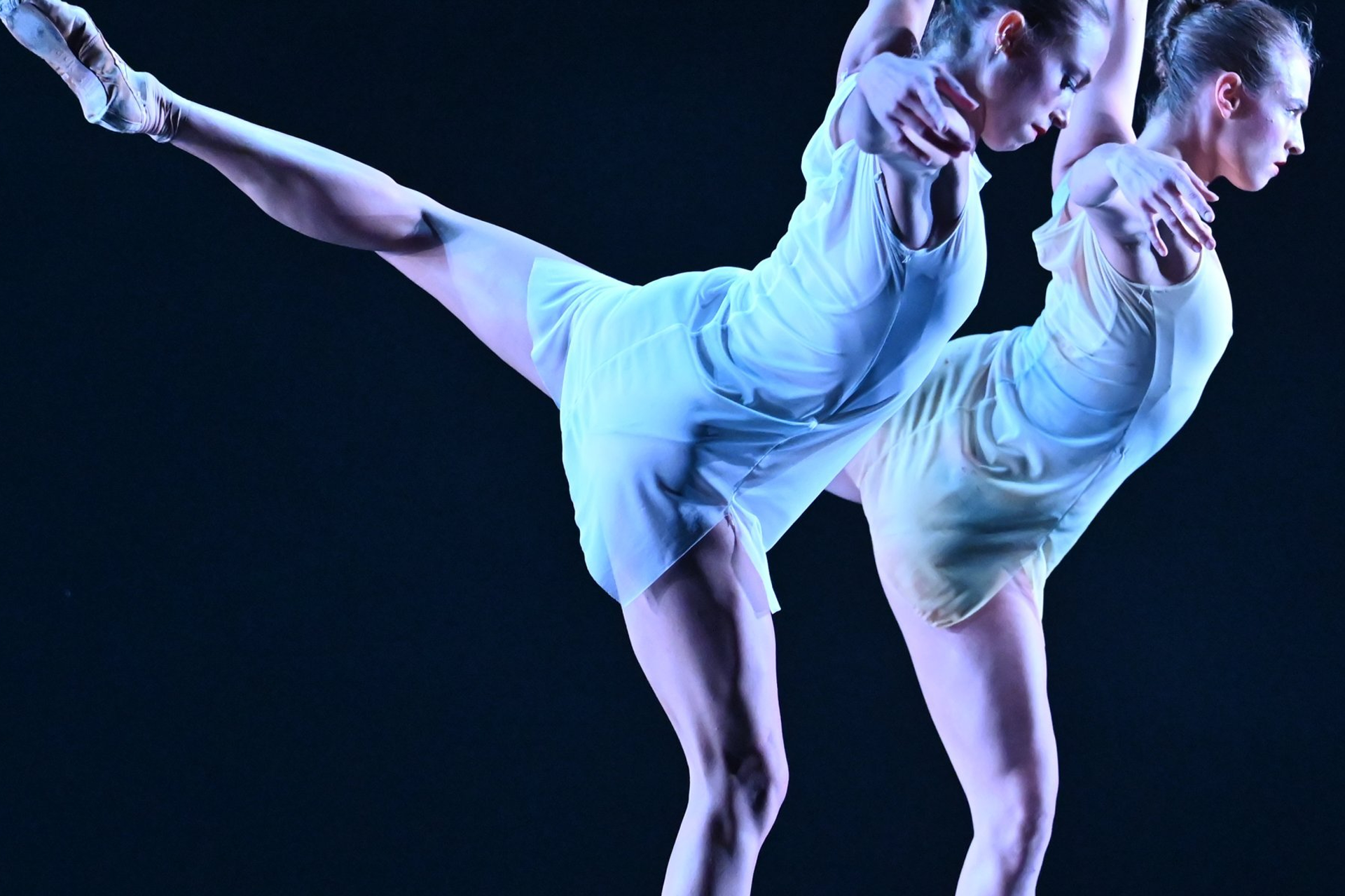 Two ballet dancers in pale blue costumes perform a mirrored pose against a dark background, each balancing on one leg with the other extended behind them, their arms linked and bodies aligned under cool stage lighting.