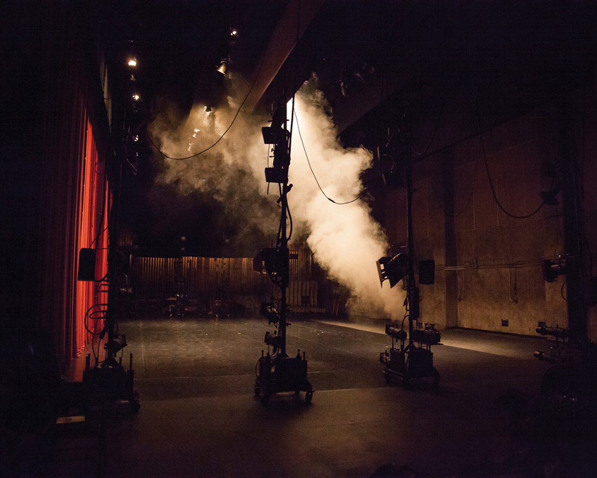 A dimly lit theater stage with red curtains on the left and stage equipment set up across the floor. Large lighting rigs and stands are visible, and a plume of theatrical smoke drifts through the space. Overhead stage lights cast a warm glow, creating a dramatic backstage atmosphere.