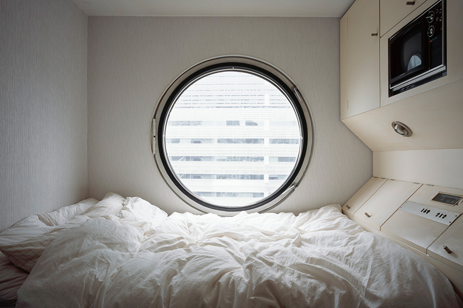 Small minimalist bedroom with a round porthole-style window centered above a rumpled white bed. Built-in white cabinets and a microwave are mounted along the wall, creating a compact, modern space with soft natural light coming through the window.